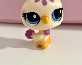 May include: A small, cream-colored bird figurine with large blue eyes, black pupils, and a yellow beak. The bird has purple accents on its head and wings, and yellow feet. This collectible toy is likely part of a larger set.