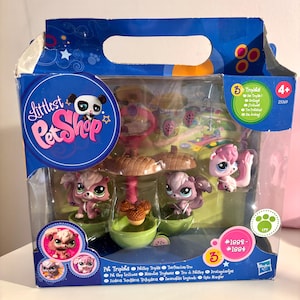 May include: A Littlest Pet Shop playset featuring three toy pets, a mushroom-shaped structure, and green bowls. The packaging is blue with the Littlest Pet Shop logo and the text "Pet Triplets". The set is designed for ages 4+.