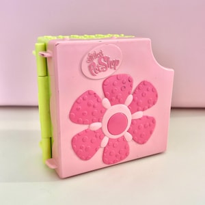 May include: A pink and green Littlest Pet Shop toy. The toy is a rectangular shape with a large pink flower design on the front. The toy has the words "Littlest Pet Shop" on the top.