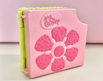 May include: A pink and green Littlest Pet Shop toy. The toy is a rectangular shape with a large pink flower design on the front. The toy has the words "Littlest Pet Shop" on the top.