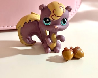 Könnte beinhalten: Eine lila-gelbe Eichhörnchenfigur mit großen blauen Augen und zwei kleinen braunen Eicheln. Das Eichhörnchen sitzt und hält einen gelben Gegenstand. Der Hintergrund ist hellrosa.