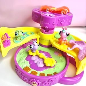 May include: A colorful toy playset featuring several small animal figurines. The playset has a purple base with a green and purple interior, yellow ramps, and a rotating top. The figurines include a green turtle, a pink insect, and a purple insect.