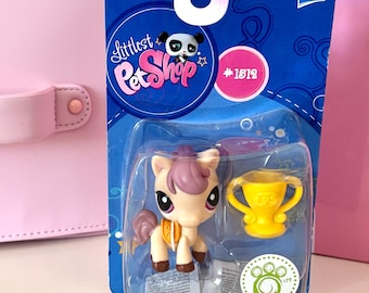 May include: A purple and white horse figurine with a yellow ribbon and a gold trophy. The figurine is part of the Littlest Pet Shop toy line. The packaging features the Littlest Pet Shop logo and the number #1512.