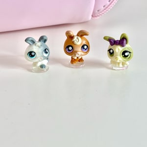 Puede incluir: Tres pequeñas figuras de conejos. Una es gris y blanca, otra marrón y la tercera blanca y verde con un lazo morado. Tienen ojos grandes y están sobre bases de plástico transparente. Una bolsa rosa en el fondo.