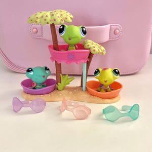 May include: A set of colorful toy frogs in a beach scene. The scene includes a pink structure with a frog, two palm trees, and two other frogs in small bowls. There are also three pairs of sunglasses in different colors.