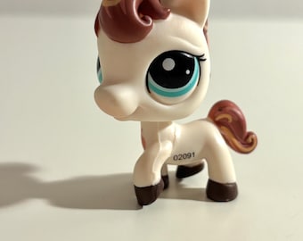 May include: A small, off-white pony figurine with brown and gold accents. The pony has large blue eyes, a brown mane, and a curly tail. The number "02091" is printed on the side. The toy is standing on all four legs.