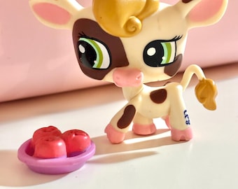 May include: A small, cream-colored cow figurine with brown spots, a brown face, and green eyes. It has a curly, golden-yellow hair tuft and pink accents on its ears and hooves. A small purple dish holds three red, heart-shaped candies.