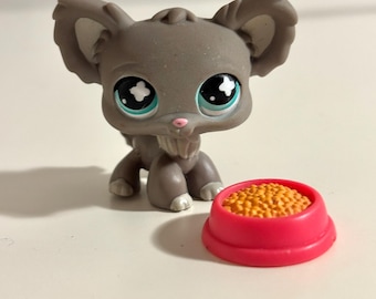 Pode incluir: Um brinquedo de animal cinzento com orelhas grandes e olhos grandes, ao lado de uma tigela rosa cheia de pellets laranja. O brinquedo tem um pequeno nariz rosa e detalhes brancos nas patas.