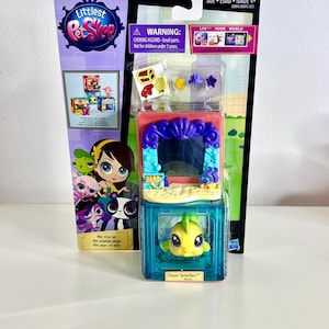 Puede incluir: Un Mini Style Set de Littlest Pet Shop con una figura amarilla de Flippa Splashley. El set incluye un juego azul y morado con accesorios. El embalaje incluye el texto "AGE 4+" y una advertencia sobre piezas pequeñas.