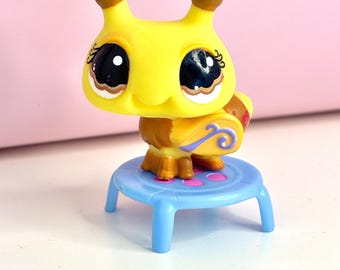 May include: A yellow and brown toy insect with large eyes and brown antennae. The toy is perched on a small, light blue plastic stool with pink dots. The background is a soft pink.