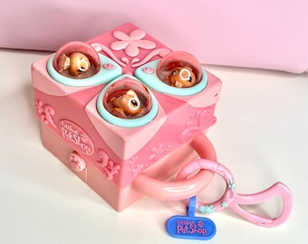 May include: A pink Littlest Pet Shop toy featuring three small animal figures in clear domes. The toy is box-shaped with a handle and a blue tag that reads "Littlest Pet Shop". Decorative floral accents are visible.