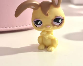 Op de afbeelding: Een gele speelgoed konijn figuur met bruine oren en accenten. Het konijn heeft grote ogen met stervormige pupillen en een kleine roze neus. Het beeldje zit in een zittende positie, met de poten tegen elkaar.