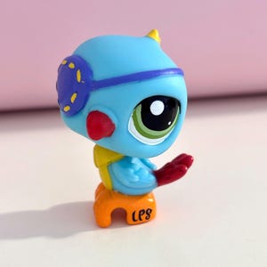 May include: A blue bird figurine with a yellow and red accents, wearing a purple eye mask. The bird is perched on an orange seat with the letters "LPS" printed on it. The bird has a large green eye.