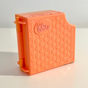 Puede incluir: Un estuche de transporte de juguetes de plástico naranja con un estampado floral y el texto "Littlest Pet Shop". El estuche es rectangular con bordes redondeados y un lado con bisagras. El estuche está cerrado.