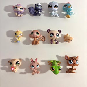 Puede incluir: Una colección de pequeñas figuras de animales de plástico de colores, incluyendo un mono, una mofeta, un pájaro, un panda y un ciervo. Cada figura tiene ojos grandes y expresivos y detalles únicos. Los juguetes están dispuestos sobre una superficie blanca.