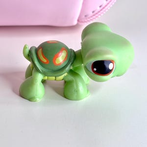 May include: A green toy turtle with a large head and big eyes. The turtle has a green shell with orange and yellow markings. The toy is on a white surface with a pink object in the background.