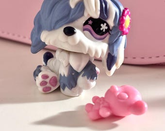 Puede incluir: Un perro de juguete blanco y azul con una flor rosa y una figura de delfín rosa. El perro tiene ojos negros y un diseño caprichoso. El pelaje del perro es blanco y azul, con detalles en rosa.