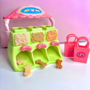 May include: A green toy pet food dispenser with a pink awning and three compartments, each with a different textured food design. Several bone-shaped treats are in front. Two pink shopping bags with "LPS" text are on the right.