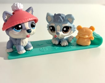 May include: Three toy figurines: two husky dogs and a small bear on a teal sled. One husky wears a red hat with a white pom-pom. The other husky and bear are orange. The sled has snowflake designs.
