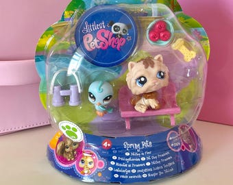 May include: A pink and blue Littlest Pet Shop playset with a small dog and bird figurine. The set includes a pink bench, a blue birdhouse, and a bowl of red berries. The packaging features the Littlest Pet Shop logo and the text "Spring Pots".
