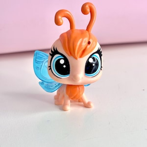 May include: A small plastic toy figure of a butterfly with a peach-colored head and body, large blue eyes with black eyelashes, and two orange antennae. The figure features translucent blue wings on its back and a small red dot on its forehead.