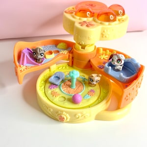 Puede incluir: Un juego de plástico amarillo y naranja con varias figuras de animales pequeños. El juego tiene múltiples niveles con varias características, incluyendo un tobogán, una piscina y una plataforma giratoria central. Las figuras incluyen un perro, un gato y un hámster.