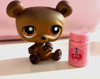 May include: A brown bear figurine with large eyes and a small pink jar with a cherry design. The bear is sitting with its paws out. The jar has a rounded top and a cherry design.