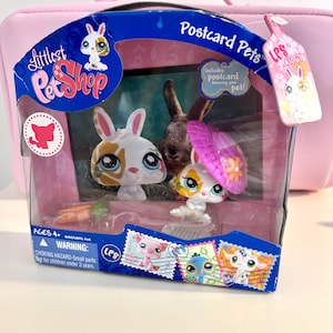 May include: A Littlest Pet Shop toy set featuring two bunny figures, one with a pink sun hat. The packaging reads "Postcard Pets" and includes a postcard. The set is in a clear plastic case with a pink background.