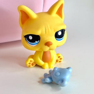 May include: A yellow cat figurine with large blue eyes and a small blue bear figurine. The cat has orange accents on its ears, nose, and paws. The cat is sitting upright, and the bear is lying down.