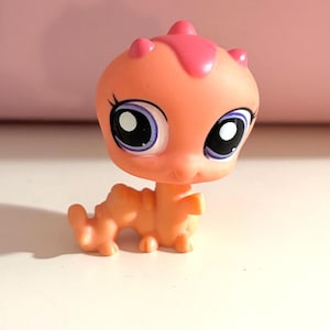 May include: A small, orange and pink toy figurine of a lizard-like creature. The figurine has large eyes with purple accents and a pink crest on its head. The toy is standing on a white surface.