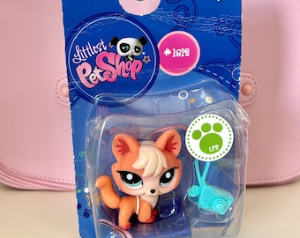 May include: A Littlest Pet Shop toy figure of a fox with orange and white fur, packaged in a clear plastic blister pack on a blue cardboard backing. The packaging includes the text "Littlest Pet Shop" and the number #1812.