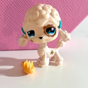 May include: A cream-coloured toy poodle figurine with a fluffy head and blue and brown eyes. The poodle has a small yellow brush beside it. The figurine has a star-shaped marking on its side.