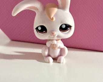 May include: A small, light pink bunny figurine with large, expressive eyes and a tuft of peach-colored fur. The bunny is in a seated position, with its paws clasped together. The background is a solid pink, and the figurine is on a white surface.