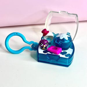 Puede incluir: Un pequeño juego de plástico azul con una tapa transparente, que presenta una figura de pájaro morado, un rectángulo rosa y un recipiente azul con un diseño de flor blanca. El juego incluye un llavero azul y un pequeño sombrero rojo.