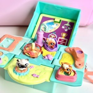 Puede incluir: Un colorido juego abierto con figuras de animales en miniatura. El juego es principalmente turquesa y amarillo, con varios compartimentos y accesorios. Varias pequeñas figuras de animales son visibles, junto con un fondo morado y rosa.