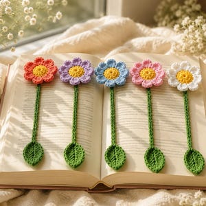 Può includere: Cinque segnalibri a forma di fiore all'uncinetto posati su un libro aperto. Ogni segnalibro presenta un lungo stelo verde con una foglia e una testa di fiore colorata con un centro giallo. I fiori sono arancioni, viola, azzurri, rosa e bianchi.