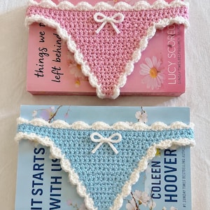 Könnte beinhalten: Zwei dreieckige Häkelkleidungsstücke, eines in Rosa und eines in Hellblau, jeweils mit weißem Muschelrand und einer kleinen weißen Schleife in der Mitte. Das rosa Stück liegt auf einem Buch namens Things We Left Behind, das blaue auf It Starts With Us.