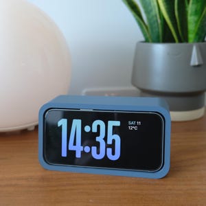 May include: A rectangular blue digital alarm clock with a black screen displaying the time 14:35 in large light blue numerals, along with the text SAT 11 and 12°C in smaller white font on the right side of the display screen.