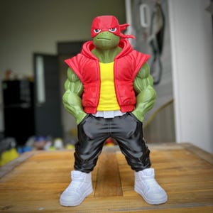 Puede incluir: Figura de acción verde de un personaje parecido a una tortuga con chaleco rojo con capucha, camisa amarilla, pantalones negros y zapatillas blancas. La figura tiene una máscara roja y posa con las manos en los bolsillos. La figura está sobre una superficie de madera.