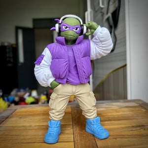 Könnte beinhalten: Eine grüne Actionfigur eines schildkrötenartigen Charakters mit einer lila Weste, weißen Ärmeln, beigefarbenen Hosen und blauen Turnschuhen. Die Figur hat eine lila Maske, weiße Kopfhörer und posiert mit einer Hand am Ohr.