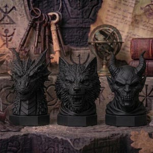 Könnte beinhalten: Drei schwarze Fantasy-Büsten: ein Drache, ein Wolf und ein gehörnter Dämon. Die Büsten stehen auf einer Steinoberfläche, mit einem Schädel, Schlüsseln und Büchern im Hintergrund. Eine brennende Kerze und ein Globus sind ebenfalls zu sehen.