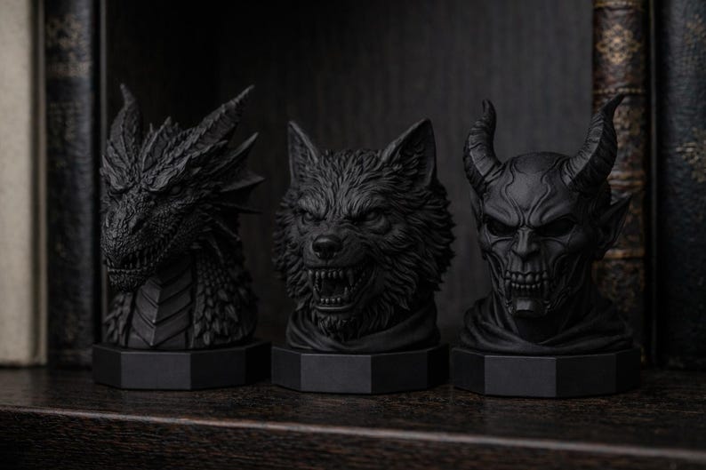 Pode incluir: Tr&ecirc;s bustos negros de fantasia: um drag&atilde;o, um lobo e um dem&ocirc;nio com chifres. Cada escultura &eacute; intrincadamente detalhada e montada em uma base hexagonal. O drag&atilde;o tem espinhos afiados, o lobo mostra os dentes e o dem&ocirc;nio tem uma express&atilde;o amea&ccedil;adora.