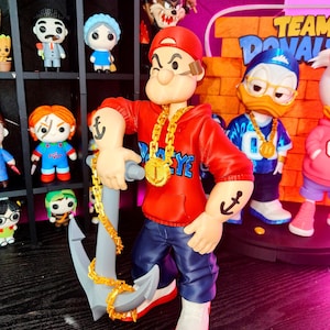 Puede incluir: Figura de Popeye con sudadera roja, vaqueros azules y zapatos blancos, sosteniendo un ancla gris con una cadena dorada. La figura tiene un collar de cadena dorada y un tatuaje de ancla. Se ven otras figuras de personajes de dibujos animados.