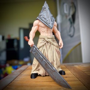 Może przedstawiać: Muskularna figurka humanoidalna w dużym, zniszczonym, trójkątnym metalowym hełmie i poszarpanej, jasnobrązowej spódnicy z materiału. Postać trzyma masywny, długi, ciemnoszary miecz z widocznymi śladami zużycia i ciemnobrązowymi detalami rękojeści.