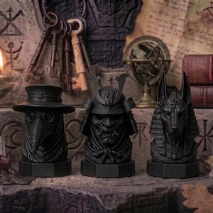 May include: Three black decorative statues: a plague doctor, a samurai, and Anubis. The statues are on a stone surface, with keys, books, and a candle in the background. The statues are approximately 6 inches tall.