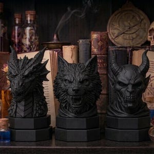 Pode incluir: Tr&ecirc;s bustos negros de fantasia: um drag&atilde;o, um lobo e um dem&ocirc;nio. Cada busto est&aacute; sobre uma base hexagonal. O fundo inclui livros, frascos de po&ccedil;&otilde;es e uma caveira, criando um tema g&oacute;tico ou de fantasia.