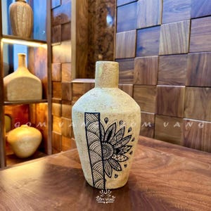 Könnte beinhalten: Eine beige Keramikvase mit einem schwarzen Blumendesign. Die Vase hat einen zylindrischen Hals und einen abgerundeten Körper. Das Blumendesign befindet sich auf der Vorderseite der Vase und umfasst eine stilisierte Blume und geometrische Muster. Die Vase steht auf einer Holzoberfläche.