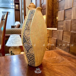 Könnte beinhalten: Eine hohe, strukturierte, hellbraune Keramikvase mit einem dekorativen schwarz-goldenen Muster auf der Vorderseite. Die Vase hat eine einzigartige Form und steht auf einer Holzoberfläche. Der Text "GOM VANG" ist sichtbar.