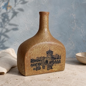 Könnte beinhalten: Eine flache, goldbraune Keramikvase mit texturierter, gesprenkelter Oberfläche und engem Hals. Die Vorderseite zeigt eine detaillierte schwarze Tuschezeichnung eines Stadtplatzes mit Uhrturm, Torbögen sowie Personen auf Motorrädern und Fahrrädern.