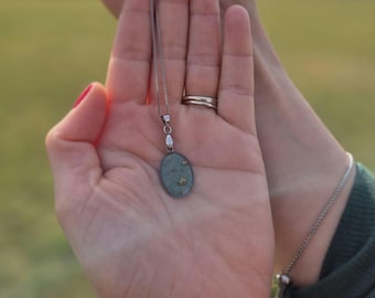 May include: A silver chain necklace with a green oval pendant containing small gold flecks. The pendant is held in a person's hand.